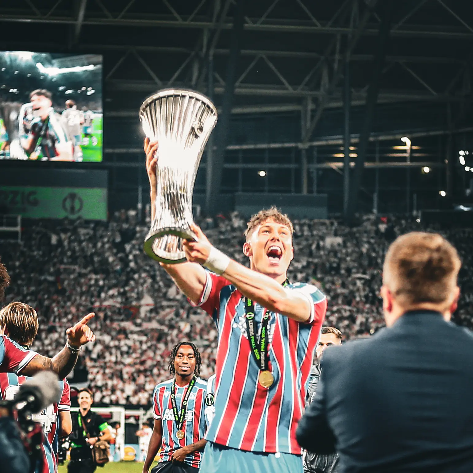 A Crystal Palace player raises the Conference League trophy toward the stands.